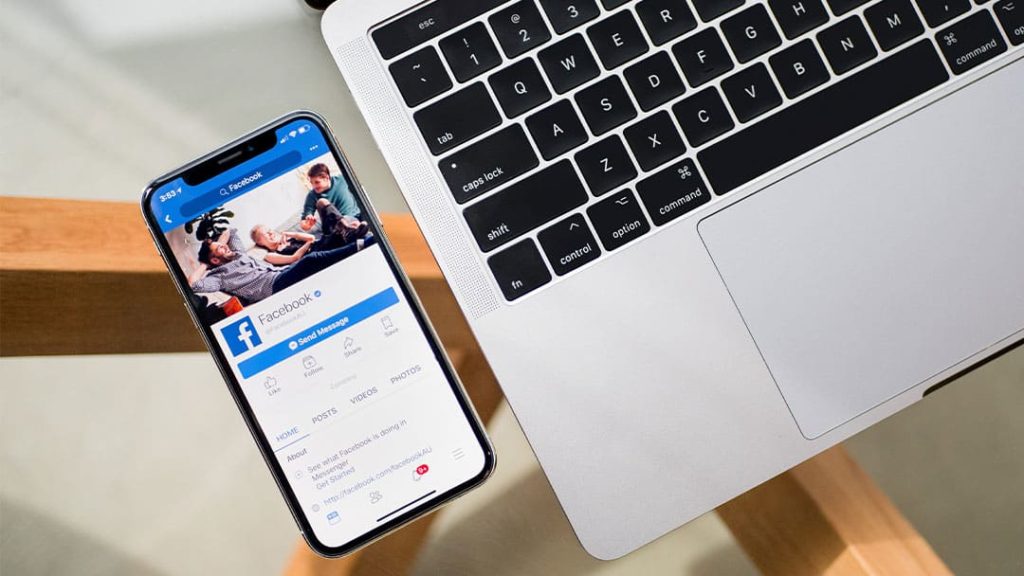 Facebook displayed on Iphone next to laptop on glass desk
